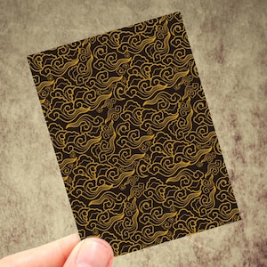 May include: A rectangular card featuring a repeating pattern of swirling gold cloud designs on a dark brown background. The card is held by a hand, showcasing the intricate details of the pattern. The design evokes a sense of traditional Asian art.