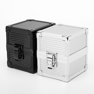 Puede incluir: Dos pequeñas cajas cuadradas con exteriores de metal acanalado. Una es negra, la otra plateada. Ambas tienen protectores de esquina de metal, una tapa con bisagras y un cierre de cierre de metal. Las cajas son probablemente para almacenamiento o exhibición.