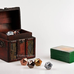 May include: A wooden treasure chest with a hinged lid, open to reveal a collection of metal dice. The chest has a brown finish with gold accents and intricate carvings. A stack of green playing cards is visible in the background.