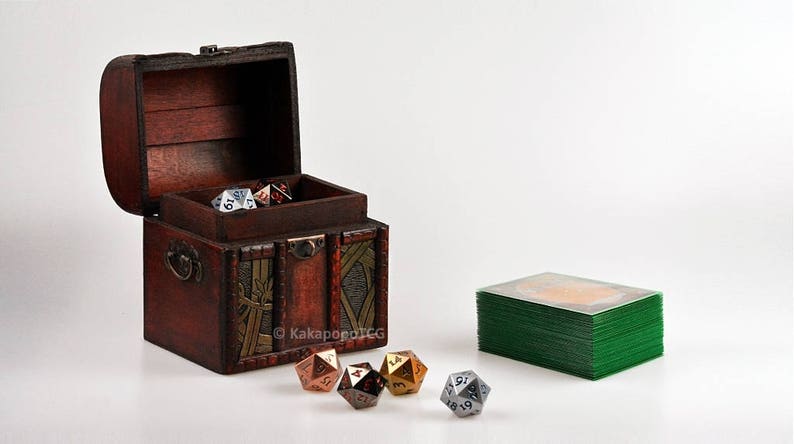 May include: A wooden treasure chest with a hinged lid, open to reveal a collection of metal dice. The chest has a brown finish with gold accents and intricate carvings. A stack of green playing cards is visible in the background.