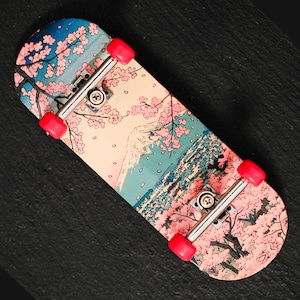 May include: A fingerboard featuring a graphic print of a mountain landscape with pink cherry blossom trees against a blue sky. The deck is equipped with silver metal trucks and four bright red wheels, showcasing a detailed artistic design.