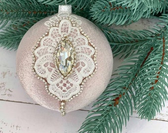 Può includere: Un ornamento natalizio in velluto rosa con un motivo in pizzo bianco e un strass trasparente al centro.