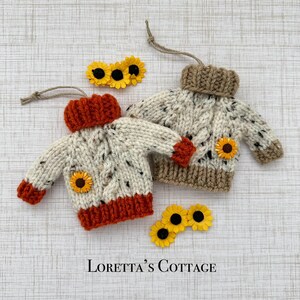 May include: Two miniature knitted sweater ornaments with hanging loops. One sweater is cream with orange trim and a sunflower accent, the other is cream with tan trim and a sunflower accent. Text at the bottom reads: Loretta's Cottage.