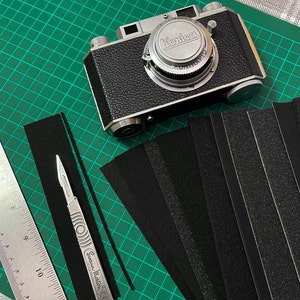 May include: A silver Konica camera with a black leather case on a green cutting mat with black strips of fabric.