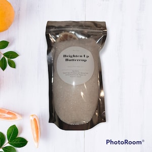 May include: A silver pouch filled with white bath salts. The pouch has a label that reads "Brighten Up Buttercup" and lists the ingredients: Epsom salt, Himalayan salt, baking soda, lemon essential oil, papaya and vitamin C serum.