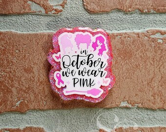 May include: A glittery pink resin badge featuring three silhouettes of women with voluminous hair, a pink awareness ribbon, small butterflies, and a broomstick. The text reads, "in October we wear PINK" in a mix of script and block lettering.