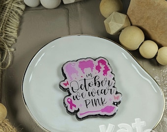 May include: A decorative acrylic pin featuring three pink silhouettes of women's hairstyles, a pink awareness ribbon, and a pink broomstick. The text reads "in October we wear PINK" in black script. The pin has a black glitter border and white base.
