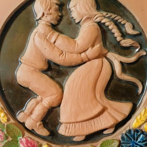 Puede incluir: Una escultura en relieve de terracota de una pareja bailando. El hombre lleva una chaqueta y pantalones marrones, y la mujer lleva un vestido largo marrón con un peinado trenzado. La escultura está rodeada por un borde floral.
