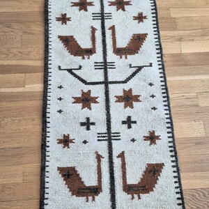 May include: A rectangular, off-white rug with a repeating pattern of brown birds, crosses, and star-like shapes. A black border frames the rug, and a central black line runs down the middle. The rug is placed on a wooden floor.