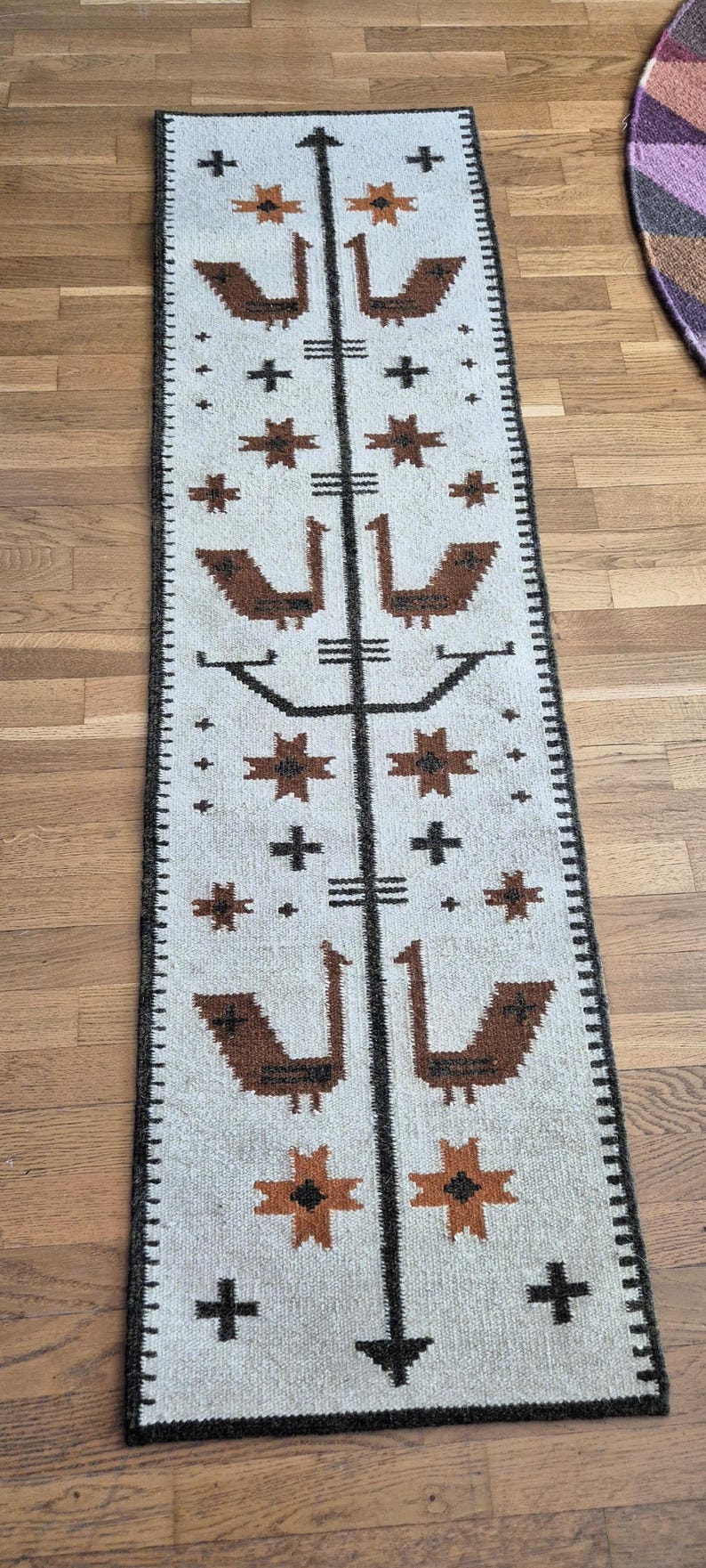 May include: A rectangular, off-white rug with a repeating pattern of brown birds, crosses, and star-like shapes. A black border frames the rug, and a central black line runs down the middle. The rug is placed on a wooden floor.