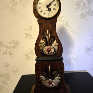 May include: A wooden clock with a painted floral design. The clock is shaped like a tall, slender vase with a round face at the top. The clock face has Roman numerals and black hands.