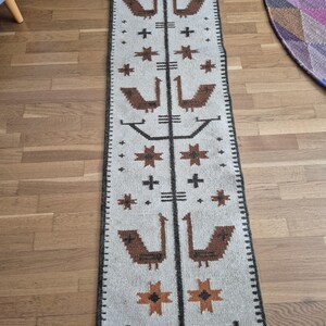May include: A rectangular, off-white rug with a repeating pattern of brown birds, crosses, and starbursts. The rug has a dark border and a central design element. The rug is approximately 60 inches long.