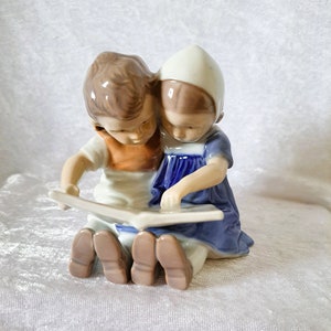 Może przedstawiać: Porcelanowa figurka przedstawiająca dwoje dzieci siedzących i czytających książkę. Dziewczynka ma na sobie niebieską sukienkę i biały czepek. Chłopiec ma na sobie brązową i pomarańczową koszulę.