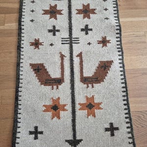 May include: A rectangular, beige-colored runner rug with a geometric pattern. The design features brown and orange star-like shapes, crosses, and bird-like figures. The rug has a dark brown border.