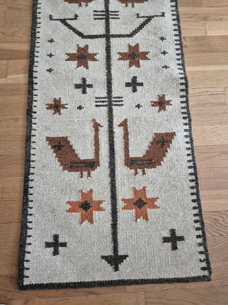 May include: A rectangular, beige-colored runner rug with a geometric pattern. The design features brown and orange star-like shapes, crosses, and bird-like figures. The rug has a dark brown border.