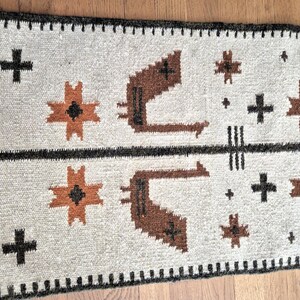 May include: A rectangular, woven rug with a cream-colored background. The rug features a black arrow design, brown geometric shapes, and black crosses. The border is black with a repeating pattern. The rug is likely made of wool.