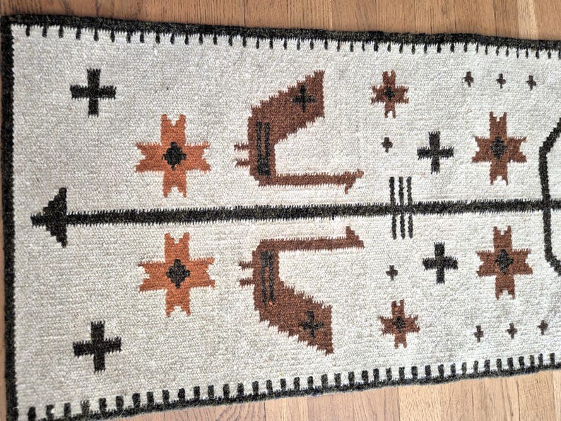 May include: A rectangular, woven rug with a cream-colored background. The rug features a black arrow design, brown geometric shapes, and black crosses. The border is black with a repeating pattern. The rug is likely made of wool.