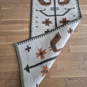 May include: A rectangular, off-white rug with a brown and black geometric pattern. The design includes stylized birds, starbursts, and cross shapes. The rug has a dark brown border and a green edge.