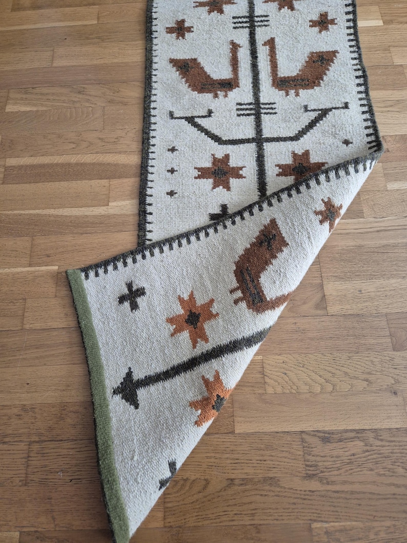 May include: A rectangular, off-white rug with a brown and black geometric pattern. The design includes stylized birds, starbursts, and cross shapes. The rug has a dark brown border and a green edge.