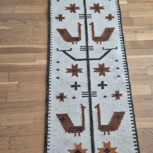 May include: A rectangular, off-white rug with a repeating pattern of brown birds, stars, and crosses. A central dark brown vertical line bisects the design. The rug has a dark brown border and is placed on a wooden floor.
