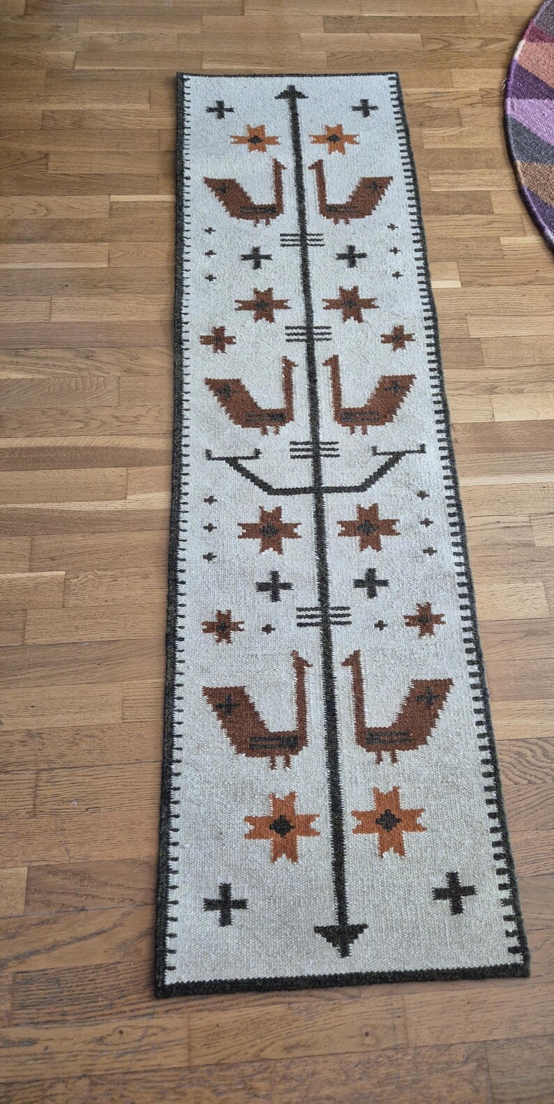 May include: A rectangular, off-white rug with a repeating pattern of brown birds, stars, and crosses. A central dark brown vertical line bisects the design. The rug has a dark brown border and is placed on a wooden floor.