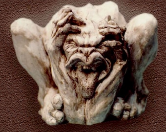 Può includere: Una scultura in ceramica bianca di una figura grottesca con le mani che si coprono il viso e la lingua che sporge.