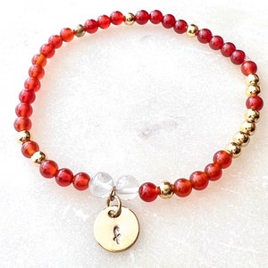 Peut inclure: Un bracelet de perles avec des perles de cornaline rouges et des accents dorés. Le bracelet comprend un charm doré avec un petit motif floral. Le bracelet est monté sur un élastique.
