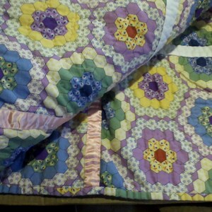 May include: A purple, blue, green, and yellow patchwork quilt with a hexagonal pattern. The quilt is folded in half and has pink satin ribbon trim.