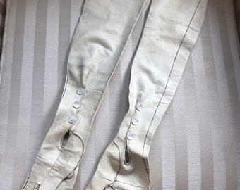 May include: A pair of long white leather gloves with pearl buttons. The gloves are laying on a striped fabric surface.