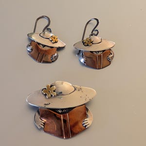 Puede incluir: Un conjunto de pendientes y un broche plateados. Cada pieza presenta una figura con un sombrero de ala ancha con una pequeña flor y sosteniendo un libro. El libro es de color cobre. Los pendientes tienen cierre de gancho.