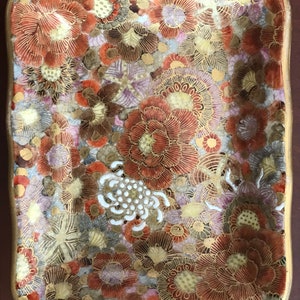 May include: A small, rectangular ceramic dish with a wavy edge. The dish is decorated with a floral pattern in shades of red, gold, and white.