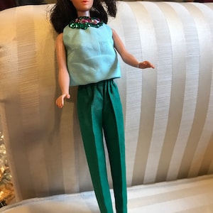 May include: A vintage doll with brown hair, wearing a light blue sleeveless top with a green and pink beaded necklace and green trousers.