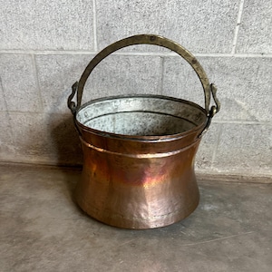 May include: A copper bucket with a brass handle. The bucket has a hammered finish and is slightly flared at the top.
