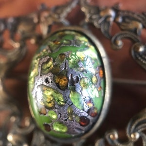 Puede incluir: Un broche vintage con un cabuj&oacute;n de esmalte verde, plateado y dorado, engastado en un marco de plata adornado.