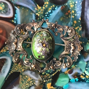 Puede incluir: Un broche vintage de forma ovalada con un centro de esmalte verde y un dise&ntilde;o filigrana intrincado en tono dorado.