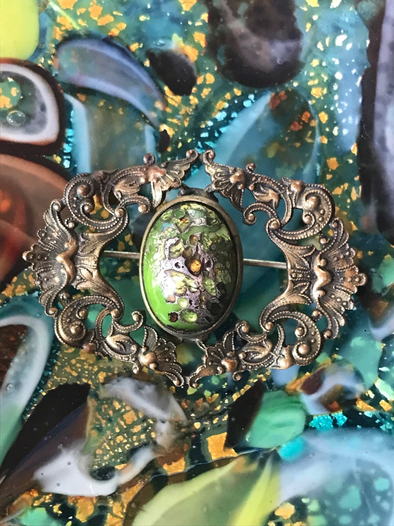 Puede incluir: Un broche vintage de forma ovalada con un centro de esmalte verde y un dise&ntilde;o filigrana intrincado en tono dorado.