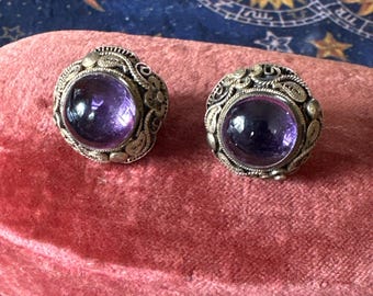 Op de afbeelding: Een paar zilveren oorbellen met paarse edelsteen-cabochons, ingelegd in ingewikkelde filigraanontwerpen.