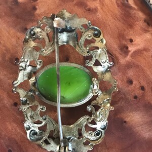 Puede incluir: Un broche de plata con un dise&ntilde;o ornamentado y un cabuj&oacute;n de vidrio verde en el centro. El broche tiene un pasador en la parte posterior para sujetarlo a la ropa.