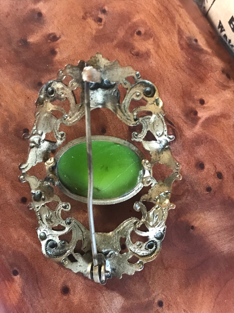 Puede incluir: Un broche de plata con un dise&ntilde;o ornamentado y un cabuj&oacute;n de vidrio verde en el centro. El broche tiene un pasador en la parte posterior para sujetarlo a la ropa.