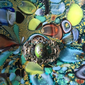Puede incluir: Un broche vintage de forma ovalada con un marco de metal dorado adornado y un centro de esmalte verde. El broche est&aacute; colocado sobre un fondo de azulejos de mosaico de vidrio de colores.