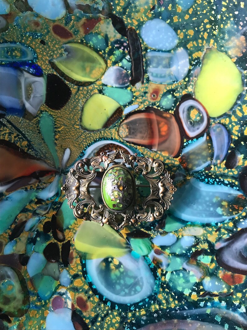 Puede incluir: Un broche vintage de forma ovalada con un marco de metal dorado adornado y un centro de esmalte verde. El broche est&aacute; colocado sobre un fondo de azulejos de mosaico de vidrio de colores.