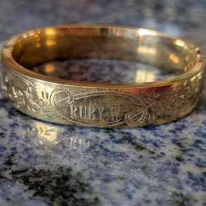 Könnte beinhalten: Ein goldfarbenes, aufklappbares Armreif-Armband mit floralen Gravuren und dem Namen "RUBY W." auf der Vorderseite. Das Armband wird auf einer blau-grau gesprenkelten Oberfläche präsentiert.