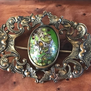 Puede incluir: Un broche antiguo con un dise&ntilde;o de esmalte verde y dorado. El broche est&aacute; hecho de metal y tiene un dise&ntilde;o de filigrana.