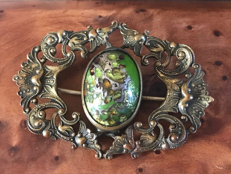 Puede incluir: Un broche antiguo con un dise&ntilde;o de esmalte verde y dorado. El broche est&aacute; hecho de metal y tiene un dise&ntilde;o de filigrana.