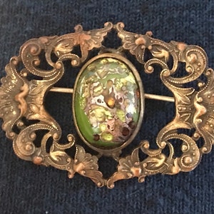 Puede incluir: Un broche vintage con un cabuj&oacute;n ovalado de vidrio verde y dorado engastado en un marco de filigrana.