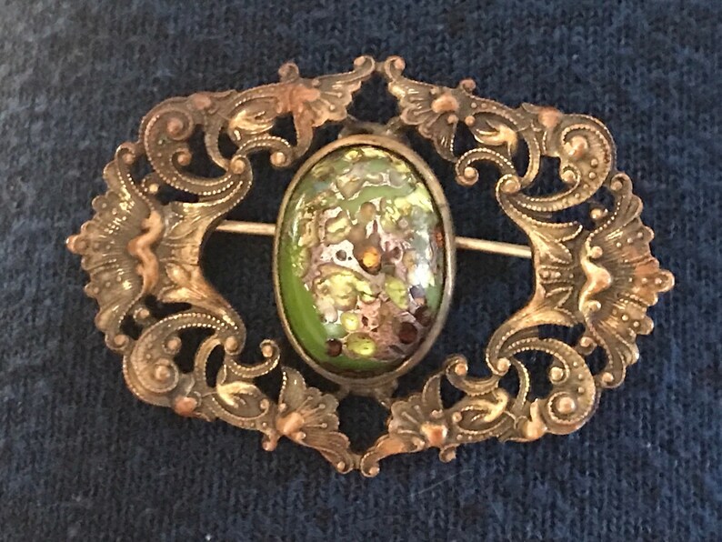 Puede incluir: Un broche vintage con un cabuj&oacute;n ovalado de vidrio verde y dorado engastado en un marco de filigrana.