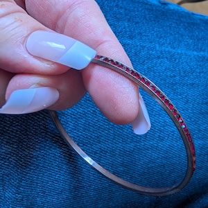 Pode incluir: Uma pulseira de bracelete prateada com uma fileira de pequenas pedras preciosas vermelhas. A pulseira é apresentada sobre um fundo de tecido de ganga azul. As pedras preciosas são uniformemente espaçadas na parte superior da pulseira, adicionando um toque de brilho.