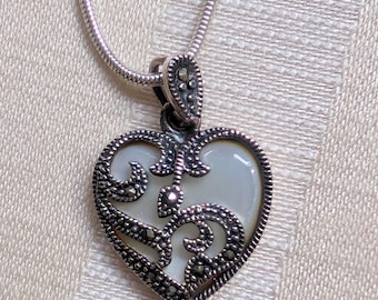 Op de afbeelding: Een zilveren ketting met een hartvormige hanger. De hanger is gemaakt van witte parelmoer en heeft een filigraanontwerp met zwarte marcasietstenen.