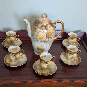 May include: A vintage tea set featuring a teapot and six teacups with saucers. The set is decorated with yellow roses and gold accents. The teapot has a curved spout and a round handle. The teacups are small and delicate.