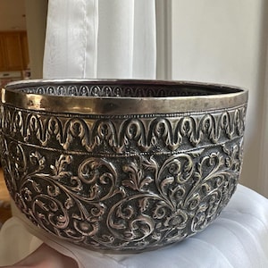 May include: A large, silver bowl with an intricate floral design. The bowl is made of metal and has a shiny finish.